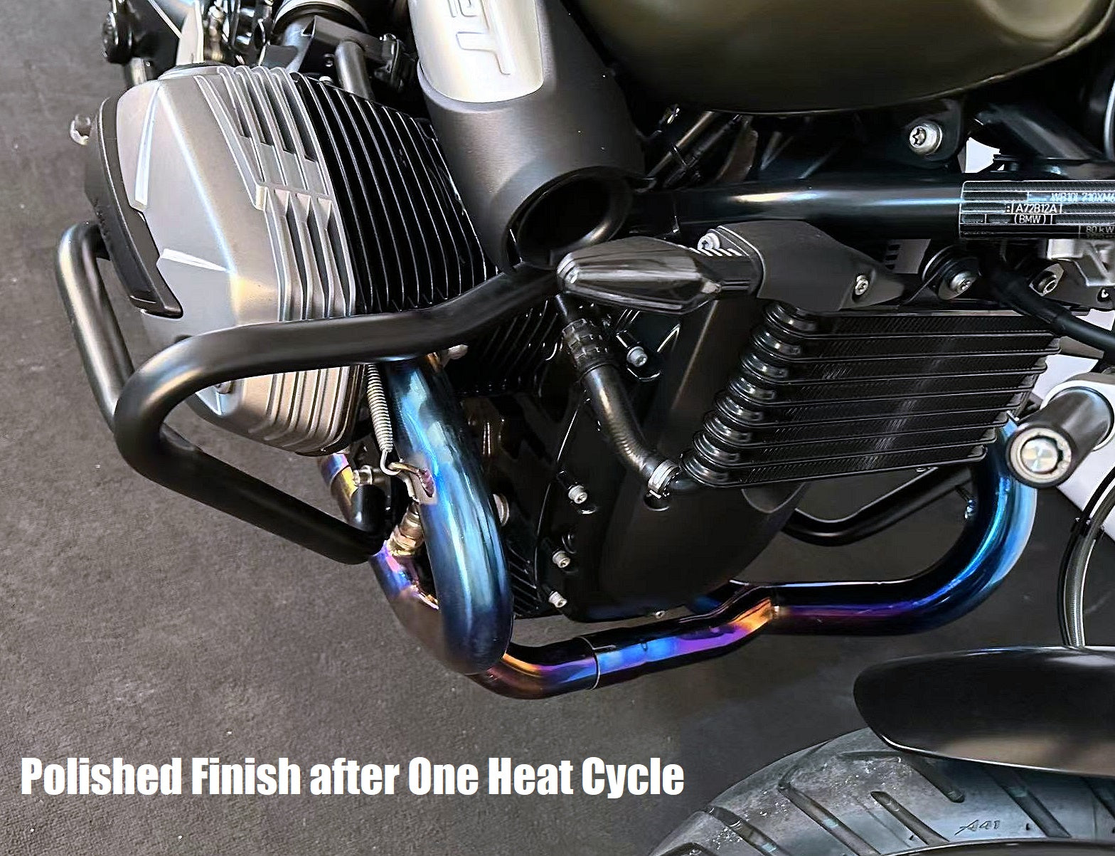 Motorcycle engine close-up showing finned cylinder head, vertically mounted oil cooler, and heat colored exhaust pipes