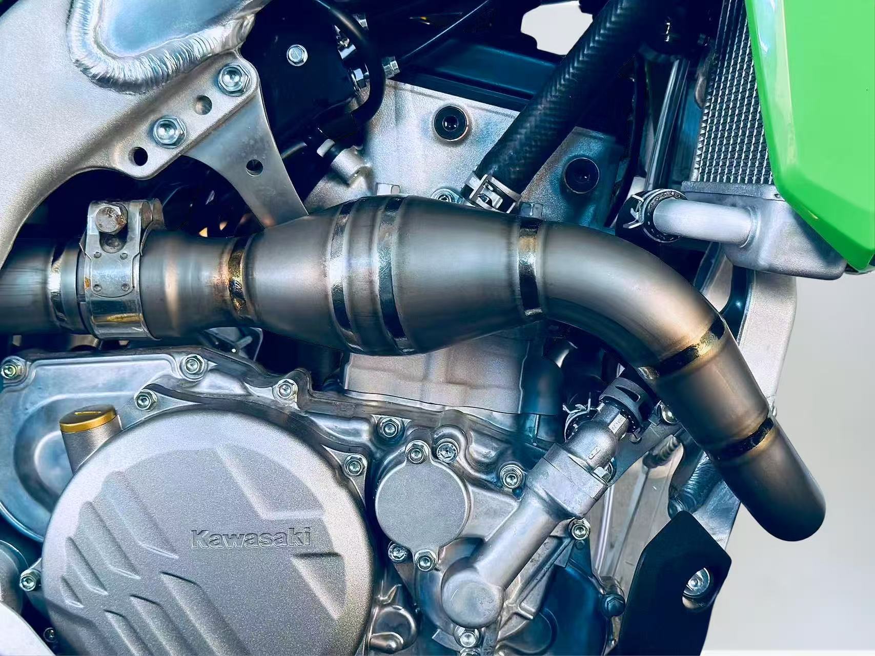 Close-up of Kawasaki motorcycle engine showing welded exhaust pipe with catalytic chamber, branded casing, and visible bolts and tubing.
