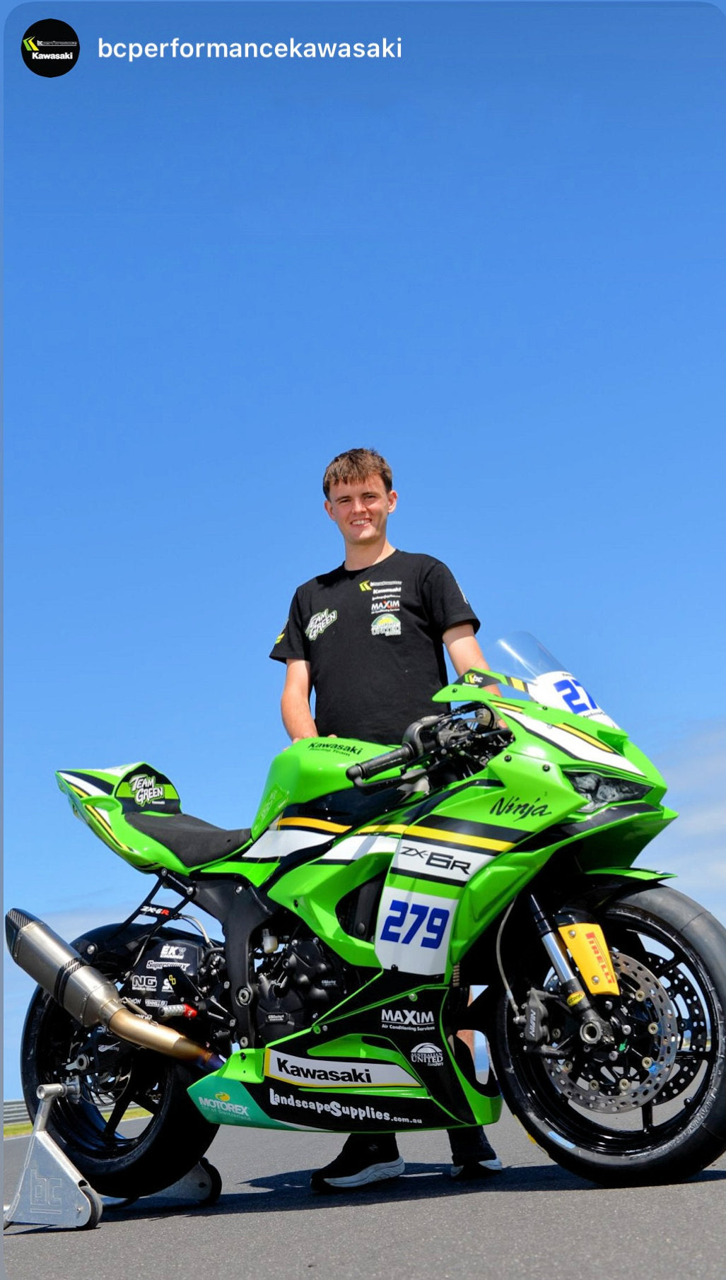 A man in black t-shirt standing beside a green motorcycle.