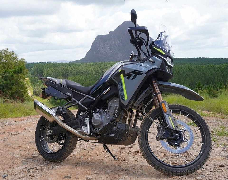 A motorcycle stands on a dirt road, surrounded by mountains, highlighting a rugged and adventurous landscape.