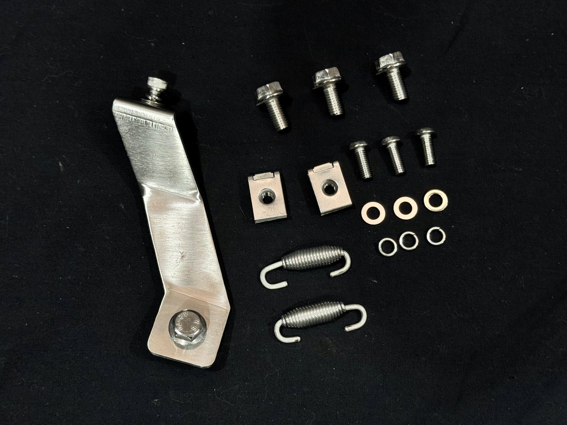 Mechanical component set on black surface including L-shaped bracket with bolt, hex head bolts, threaded plates, tension springs, washers, and spacers.