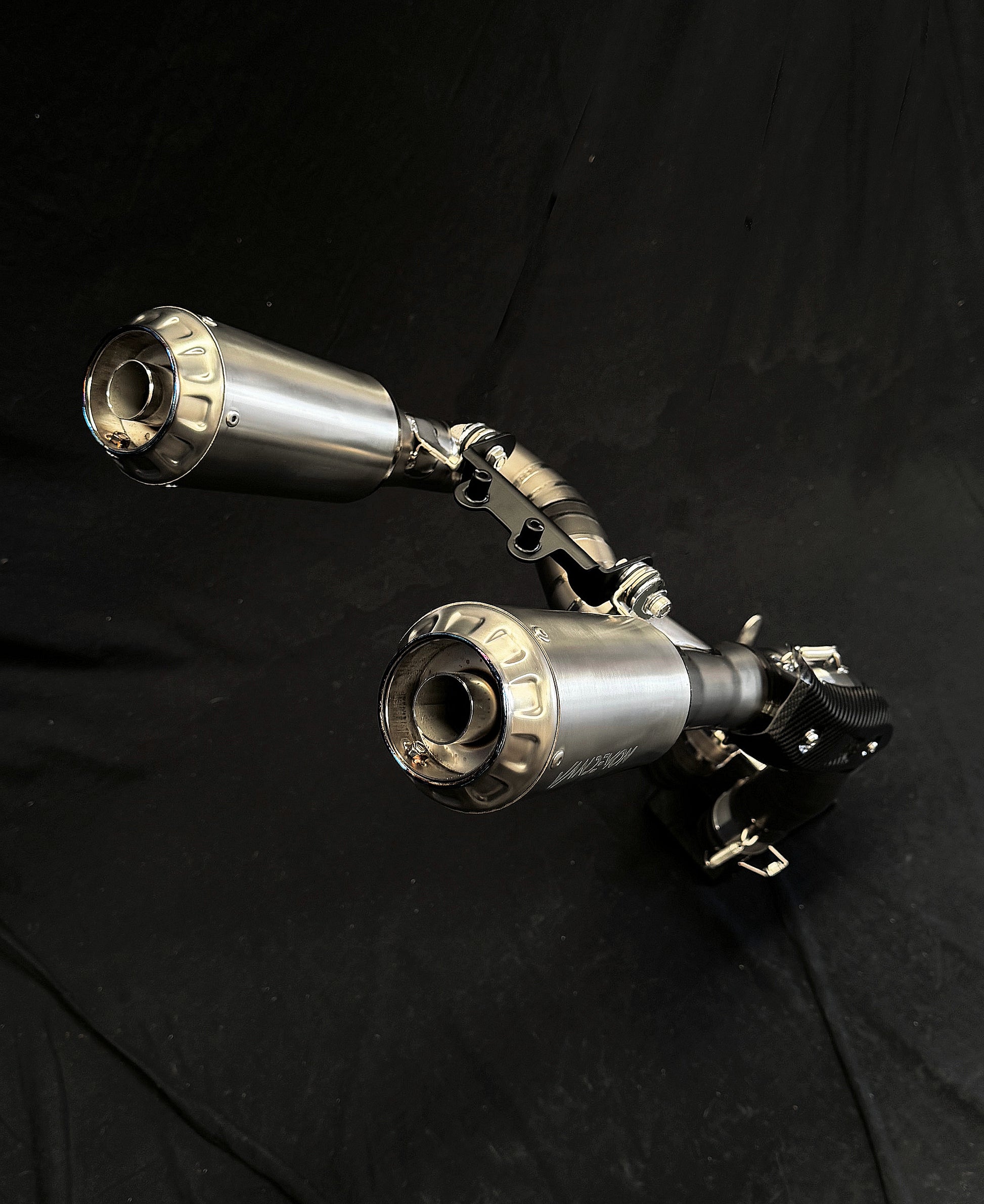Close-up of a sleek, dual-exhaust motorcycle exhaust system against a dark background. The metallic finish reflects light, highlighting its modern design and intricate detailing.