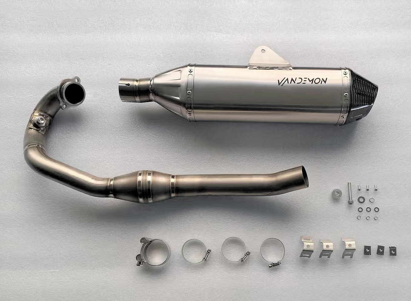 motorcycle exhaust kit laid out on a surface, showing a stainless muffler with carbon cap, curved pipe, and mounting hardware.