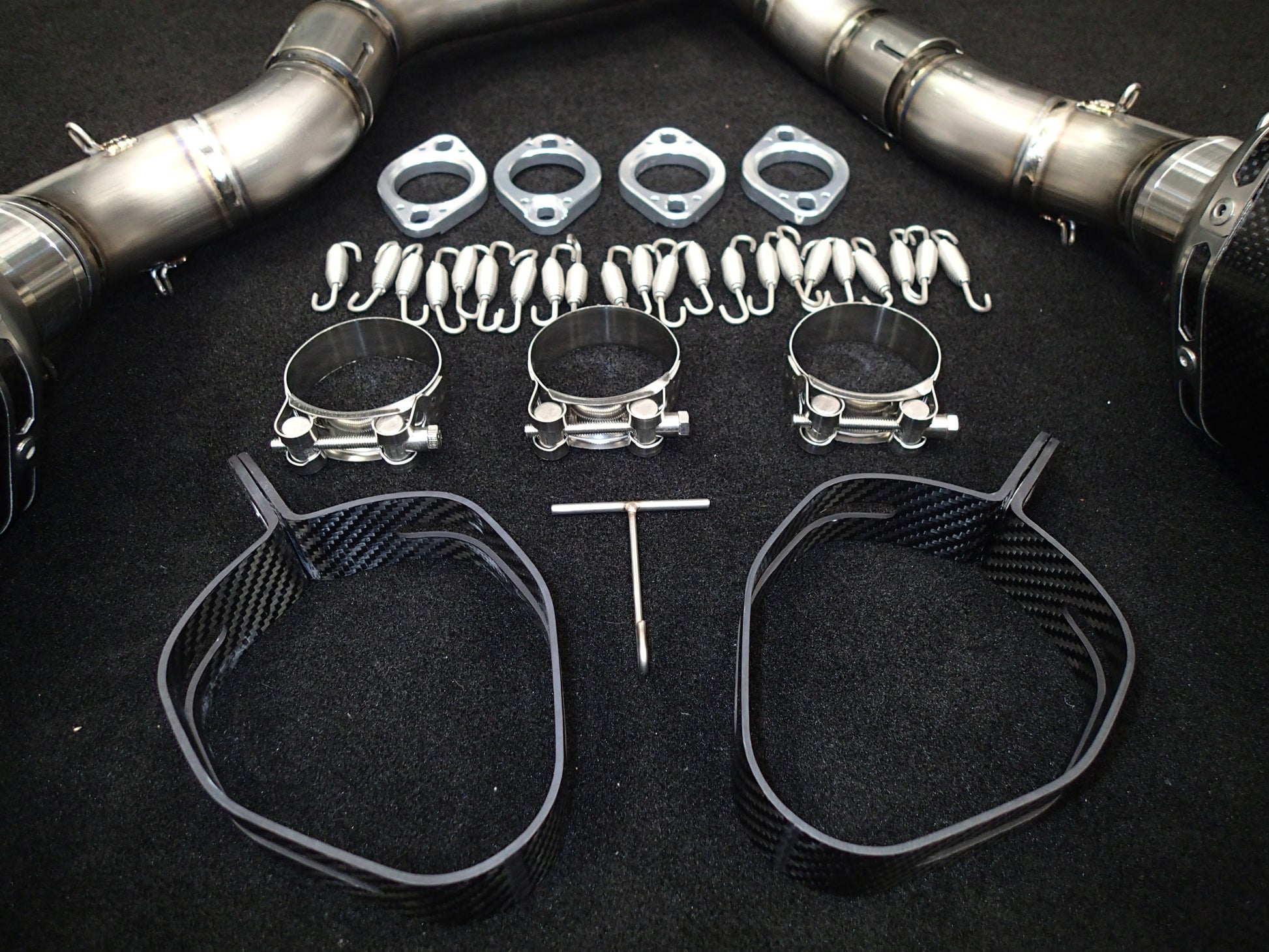 Automotive exhaust system kit on black surface featuring two curved pipes with flanges and heat shields, circular clamps, metal gaskets, spring hooks, carbon fiber hanger straps, and a T-handle hex key.