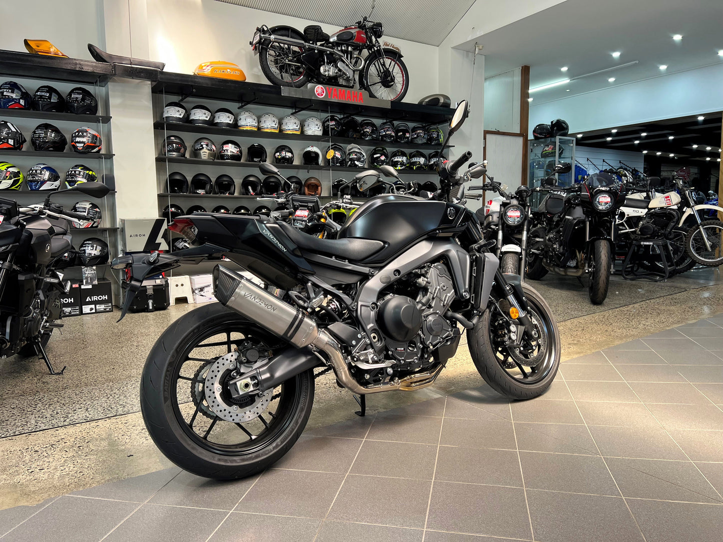 Grey motorcycle inside shop, helmets visible in background.