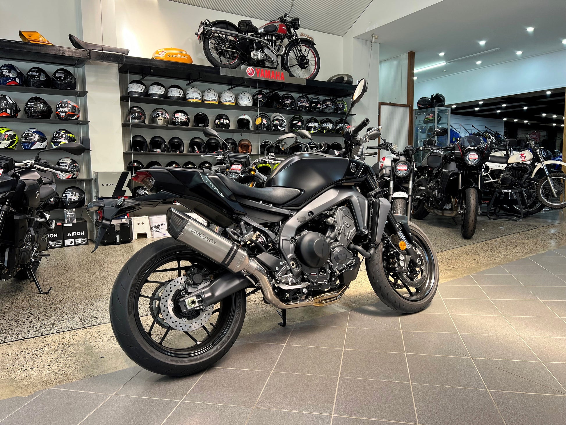 Grey motorcycle inside shop, helmets visible in background.