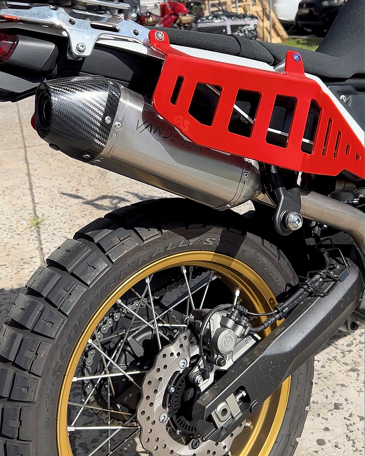 Close-up of a motorcycles rear section showing a silver exhaust with a carbon fiber tip, red accents, and gold-rimmed tires.