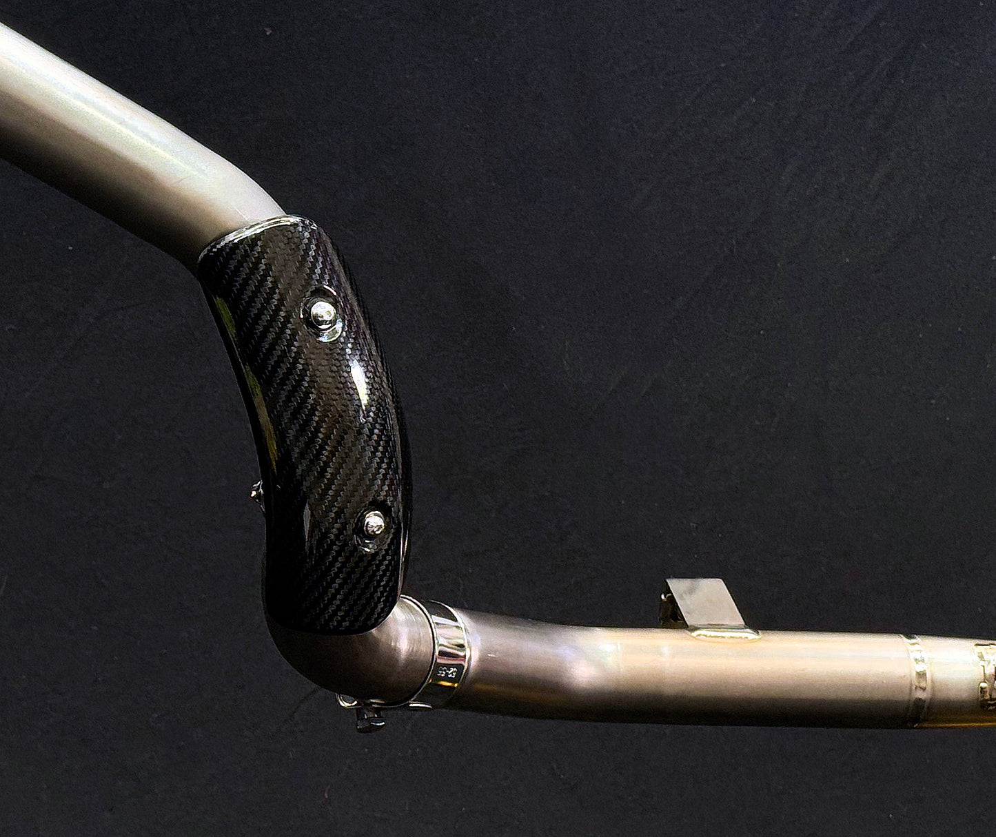 Metallic pipe with carbon fiber heat shield secured by screws, featuring a mounting bracket and braided support cables.