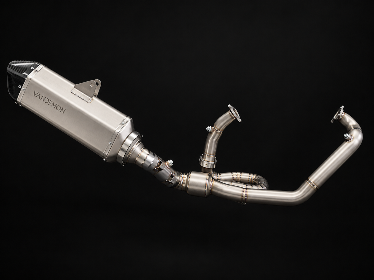 A Vandemon motorcycle exhaust system with polished stainless steel pipes and a muffler, shown against a solid black background.