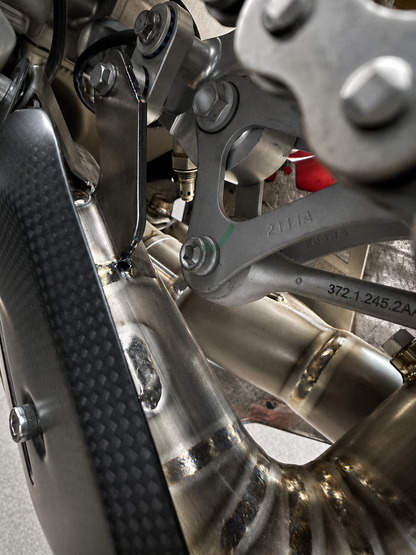 Close-up of a motorcycle's metallic exhaust system and suspension components, including a carbon fiber side panel and silver metal pipes.