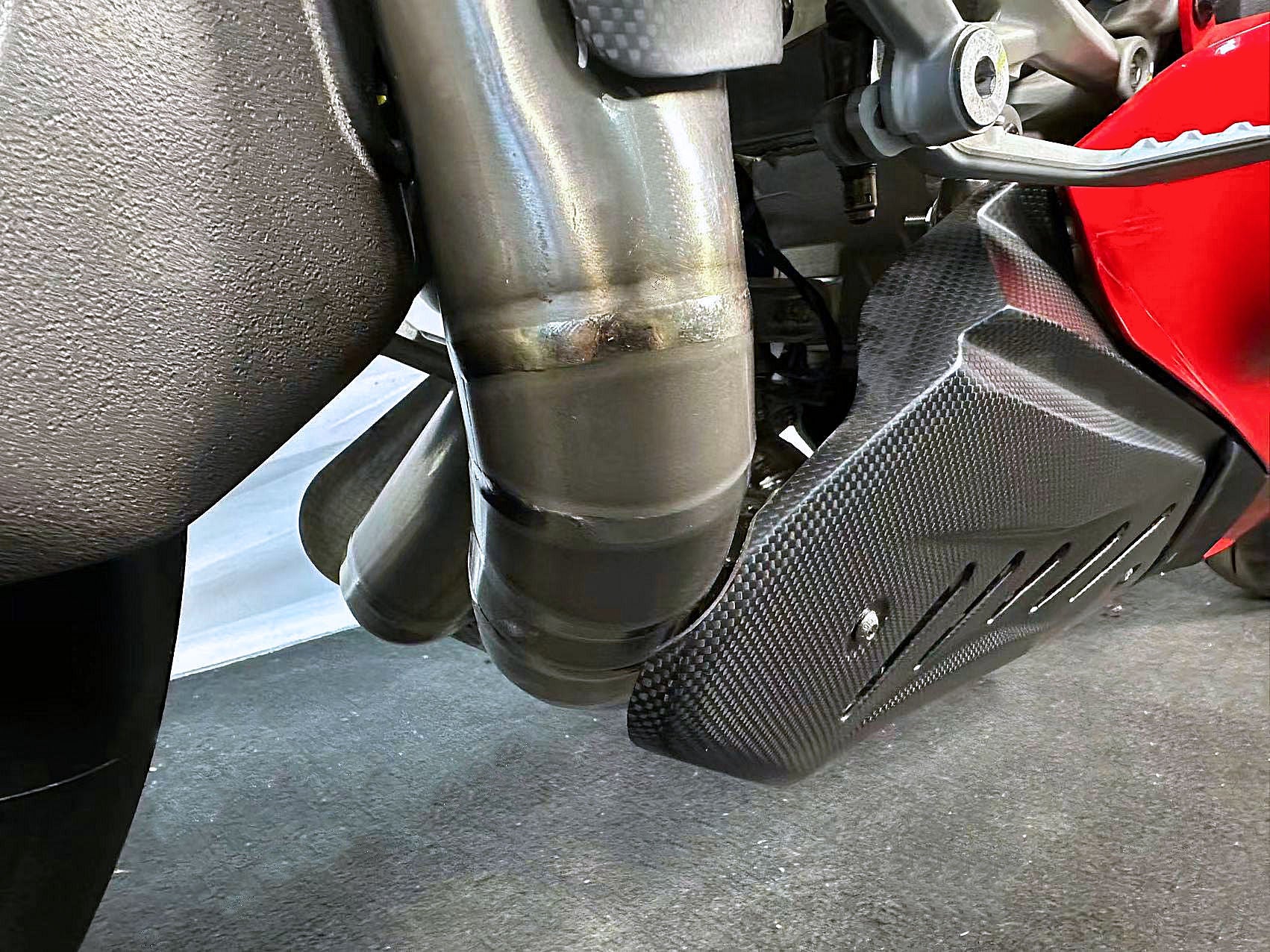 Close-up of a motorcycle exhaust and carbon fiber foot pegs on a textured surface.