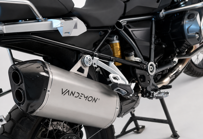 A close-up studio shot of a motorcycle's exhaust system, featuring a prominent silver and black muffler mounted on the side of a black and silver bike.
