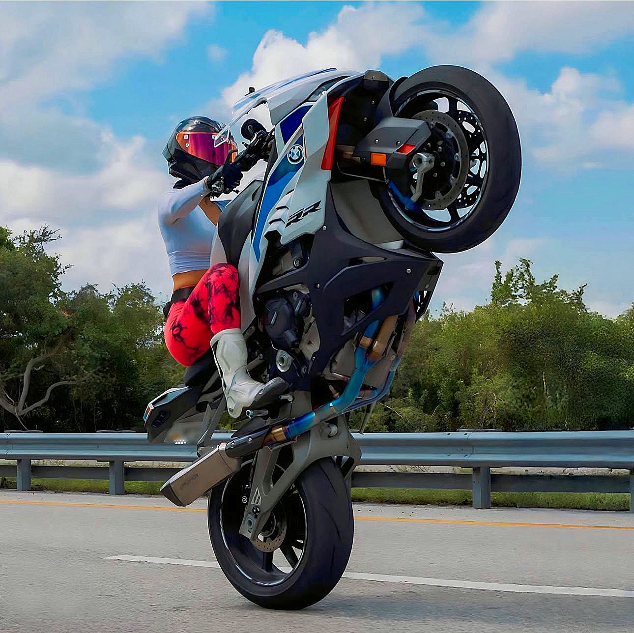 Motorcyclist performing a wheelie on a road with trees and blue sky in the background