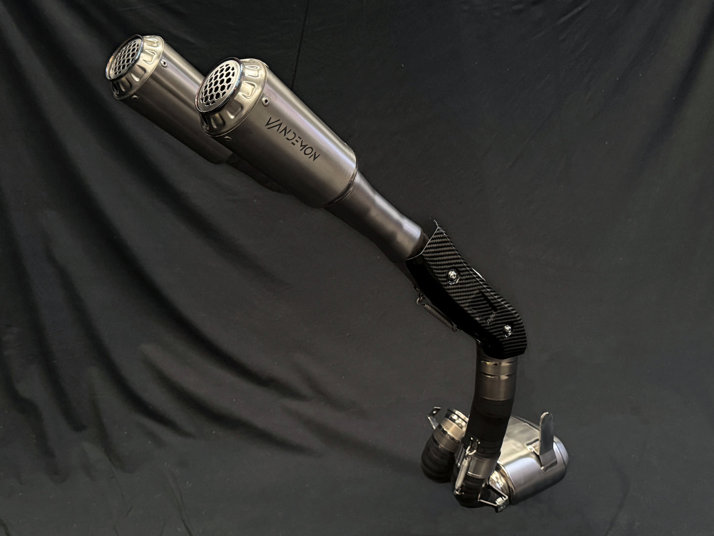 A pair of metallic exhaust pipes with a carbon-fiber joint, positioned vertically against a dark background.