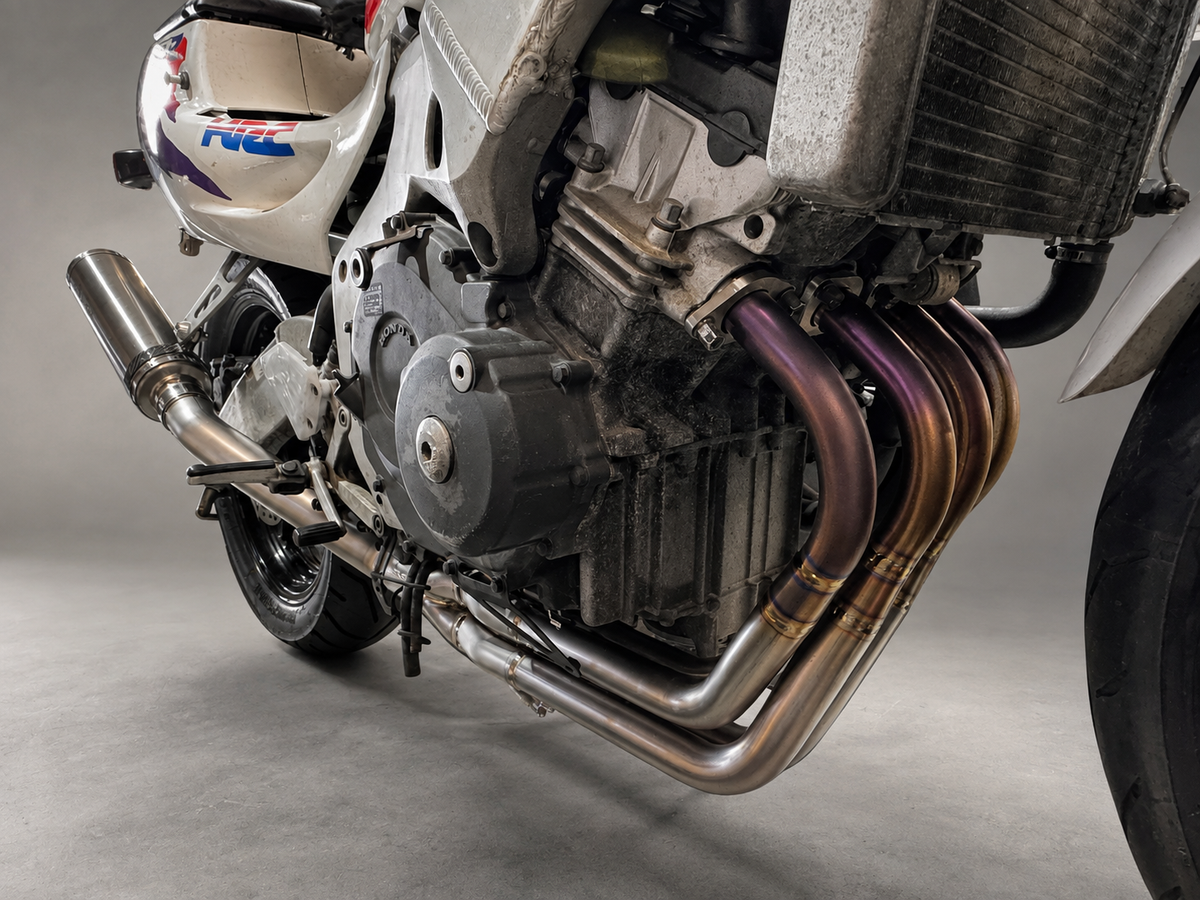 Close-up of a motorcycle engine and exhaust system, showing a polished stainless steel dual exhaust pipe with purple heat-tinted sections and a silver muffler.