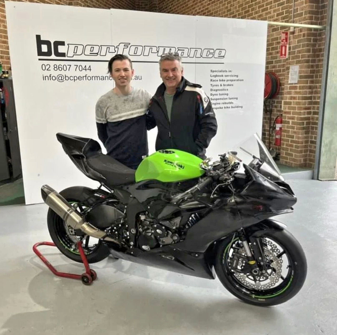 Two men standing next to a green and black motorcycle in front of a 'bc performance' banner.