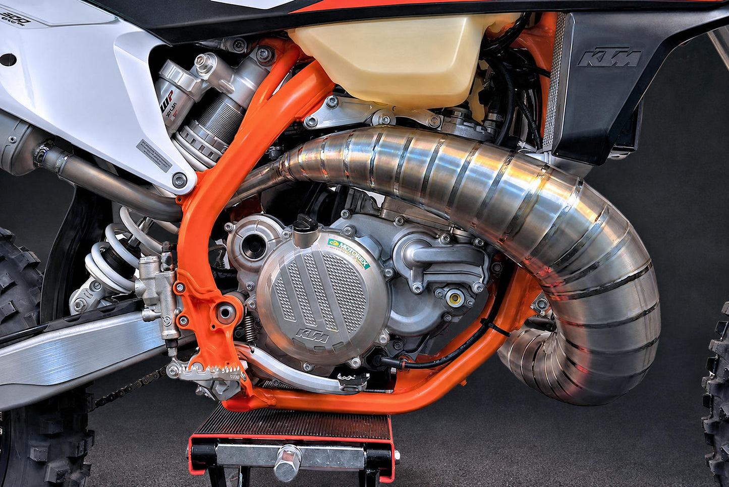 Close-up of a KTM dirt bike's engine and exhaust system, featuring a prominent silver ribbed metal exhaust pipe, a bright orange frame, and a silver engine cover.