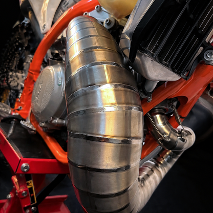 A close-up of a custom-welded stainless steel exhaust pipe mounted on an orange motorcycle engine.